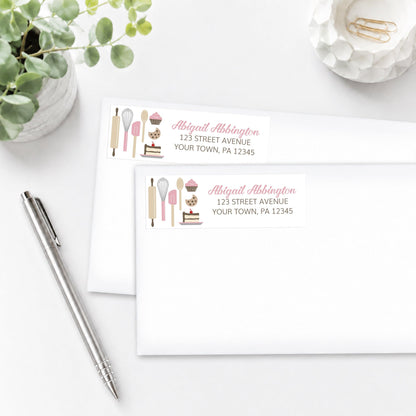 Pink Baking Address Labels with baking-themed design, shown on white envelopes, on a white surface with a pen and plant.