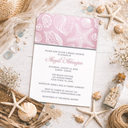 Pink Beach Seashell Pattern Bridal Shower Invitation shown on a beachy surface with sand and coastal decor.