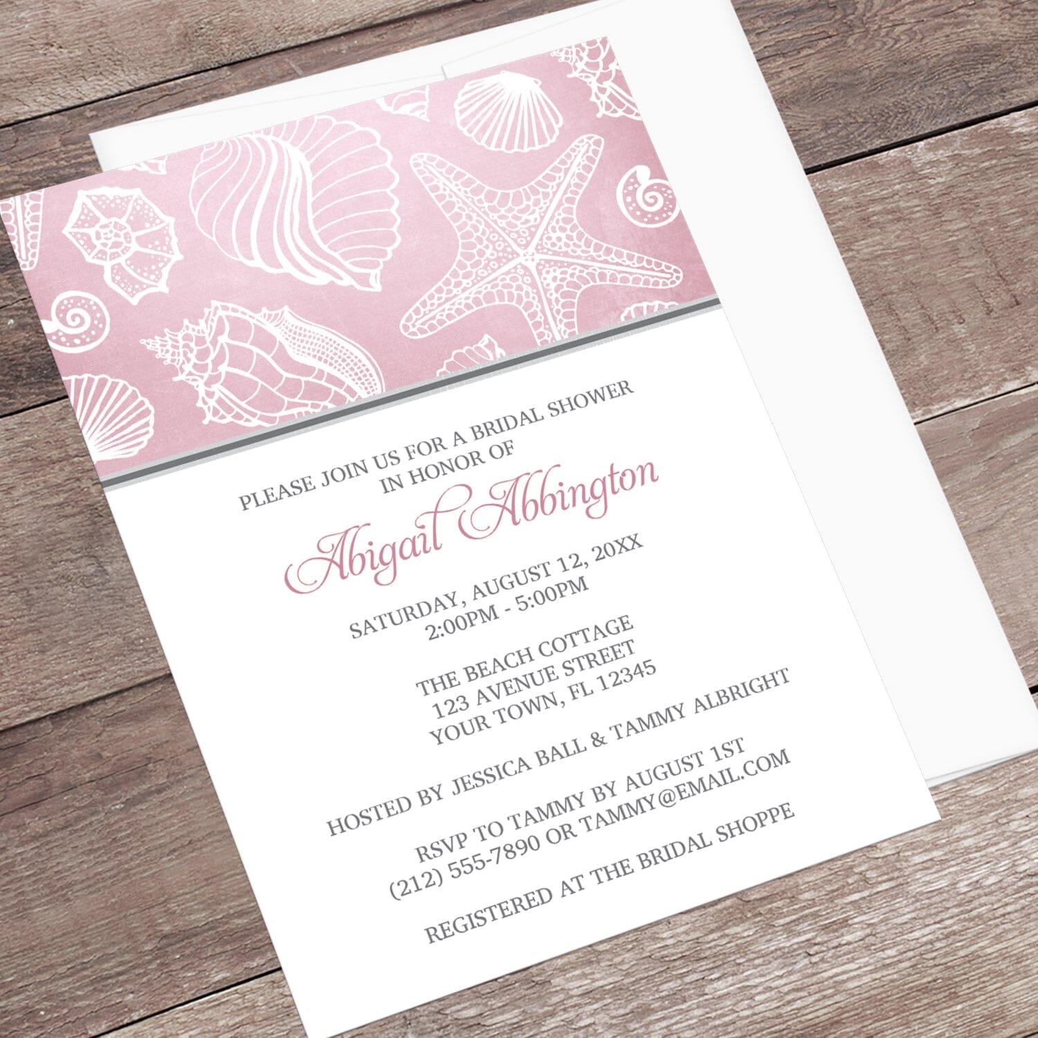 Pink Beach Seashell Pattern Bridal Shower Invitation shown at an angle on a wooden surface with a white envelope.