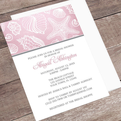 Pink Beach Seashell Pattern Bridal Shower Invitation shown at an angle on a wooden surface with a white envelope.