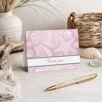 Pink Beach Seashells Pattern Thank You Card shown on a beach house tabletop with coastal decor and a pen.