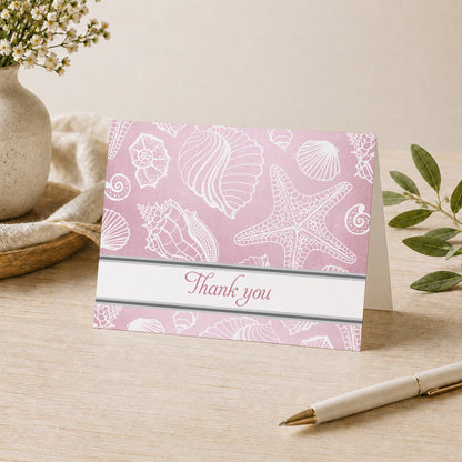 Pink Beach Seashells Pattern Thank You Card shown on a desk with a vase, plants, and a pen.