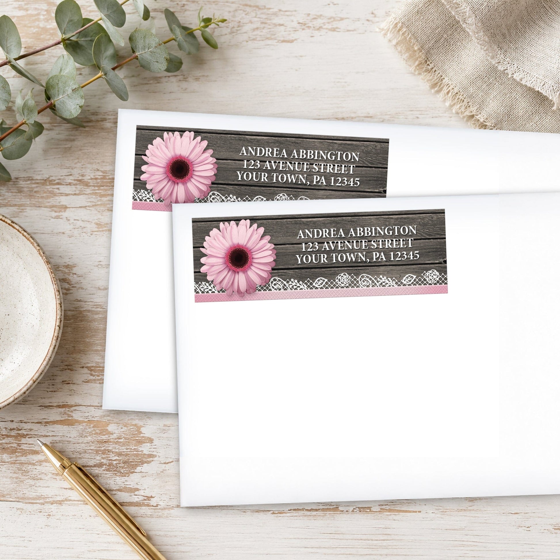 Address labels with pink daisies, lace, and wood design on white envelopes, placed on a wooden surface with plants and a pen.