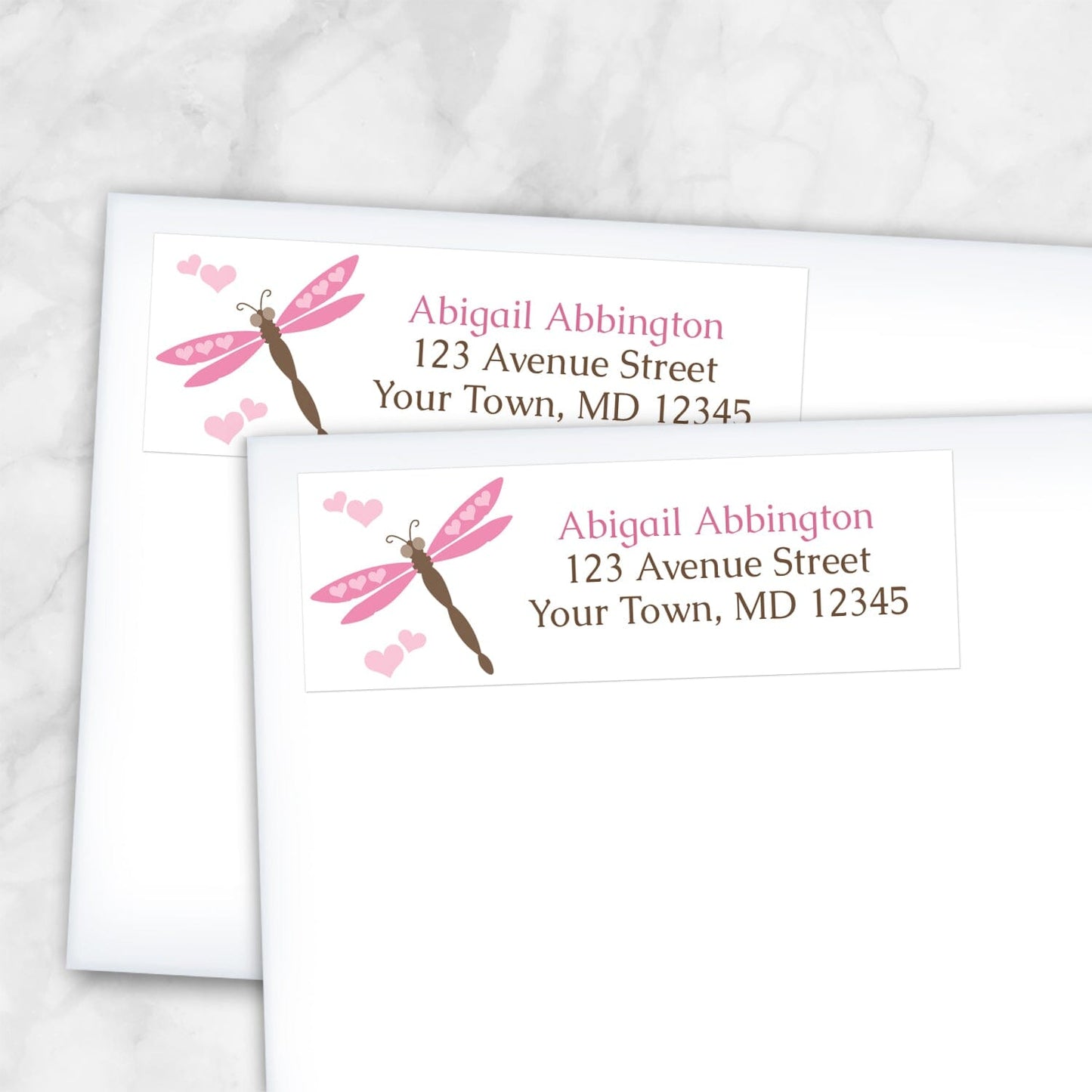 Two personalized Pink Dragonfly Address Labels with a pink and brown dragonfly and pink hearts, shown on white envelopes on a marble surface.