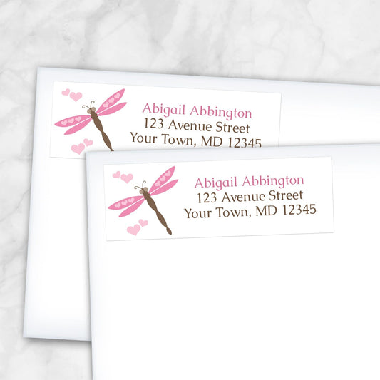 Two personalized Pink Dragonfly Address Labels with a pink and brown dragonfly and pink hearts, shown on white envelopes on a marble surface.