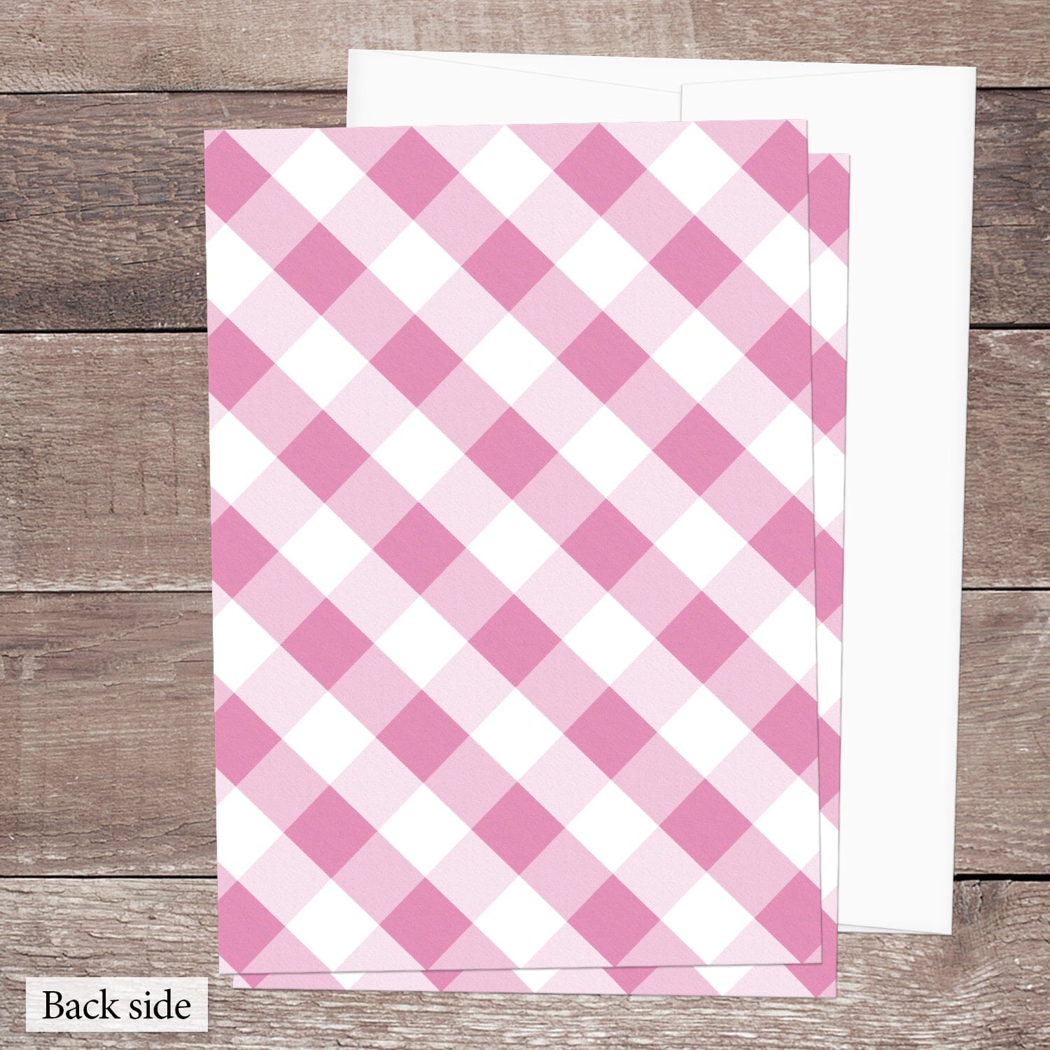 The back side of the Pink Gingham Vow Renewal Invitation, featuring the diagonal pink and white check pattern, shown on a brown wood surface with a white envelope.
