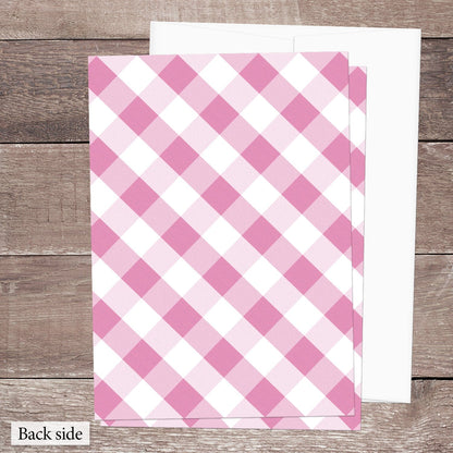 The back side of the Pink Gingham Vow Renewal Invitation, featuring the diagonal pink and white check pattern, shown on a brown wood surface with a white envelope.