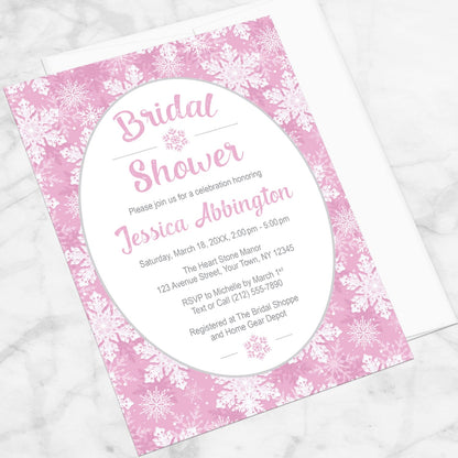 Pink Snowflake Bridal Shower Invitation shown at an angle on a marble surface with a white envelope.