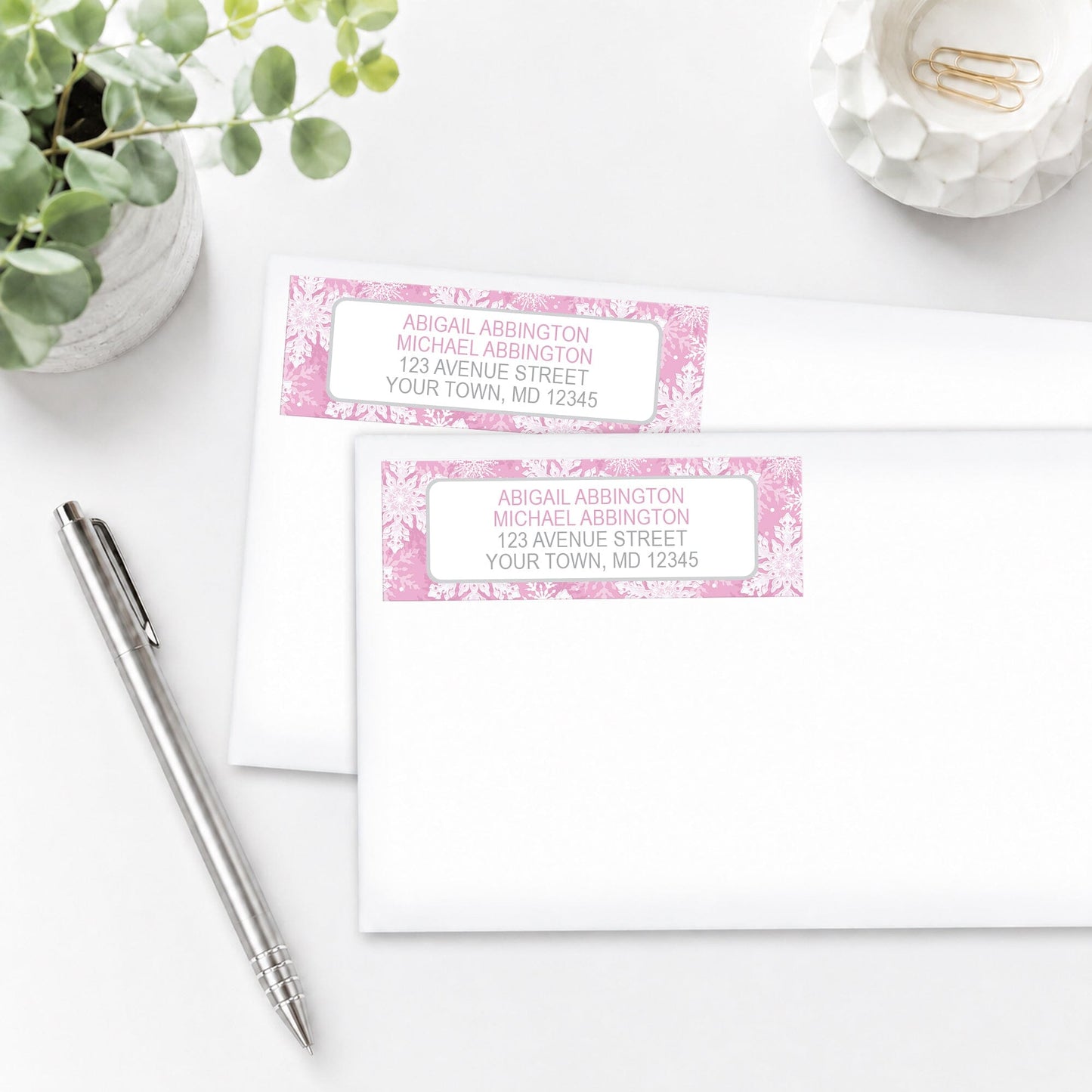 Pink Snowflake Winter Address Labels on white envelopes, shown on a white surface with a plant and a pen.