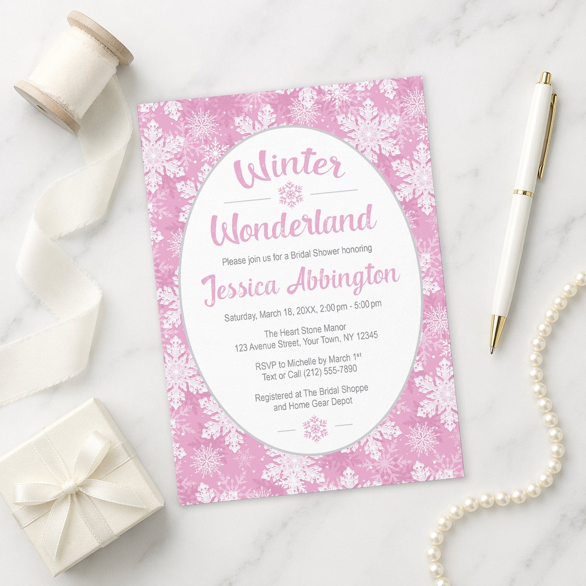 Pink Snowflake Winter Wonderland Bridal Shower Invitation shown on a marble surface with a white gift box, pen, ribbon, and pearls.