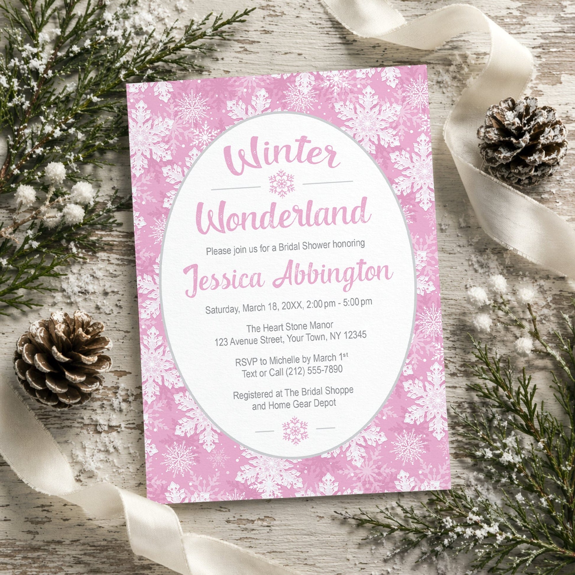 Pink Snowflake Winter Wonderland Bridal Shower Invitation shown on a rustic wooden surface with winter greenery, pine cones, and ribbons.