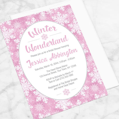 Pink Snowflake Winter Wonderland Bridal Shower Invitation shown at an angle on a marble surface with a white envelope.