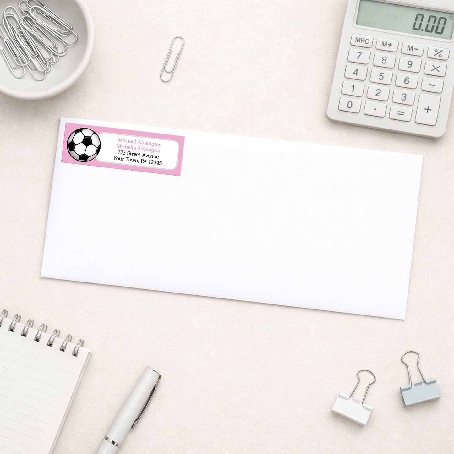 Pink Soccer Ball Return Address Label shown on a white envelope with everyday office supplies