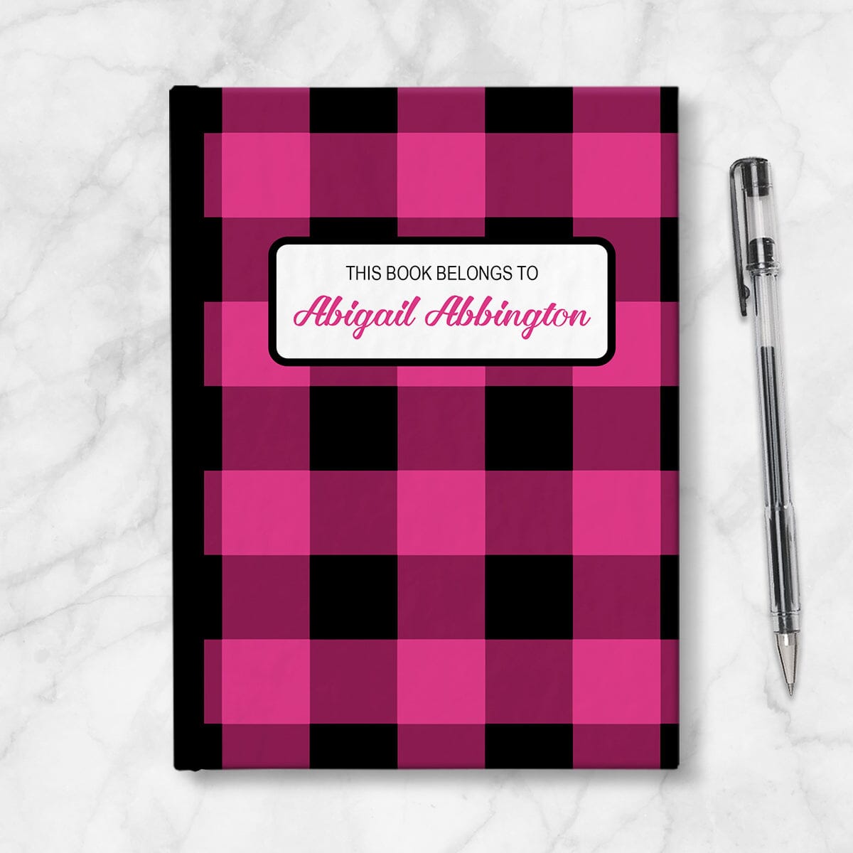 Personalized Pink and Black Buffalo Plaid Journal at Artistically Invited. Image shows the book on a countertop next to a pen.