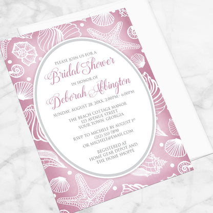 Pretty Pink Beach Seashell Bridal Shower Invitation shown at an angle on a marble surface with a white envelope.