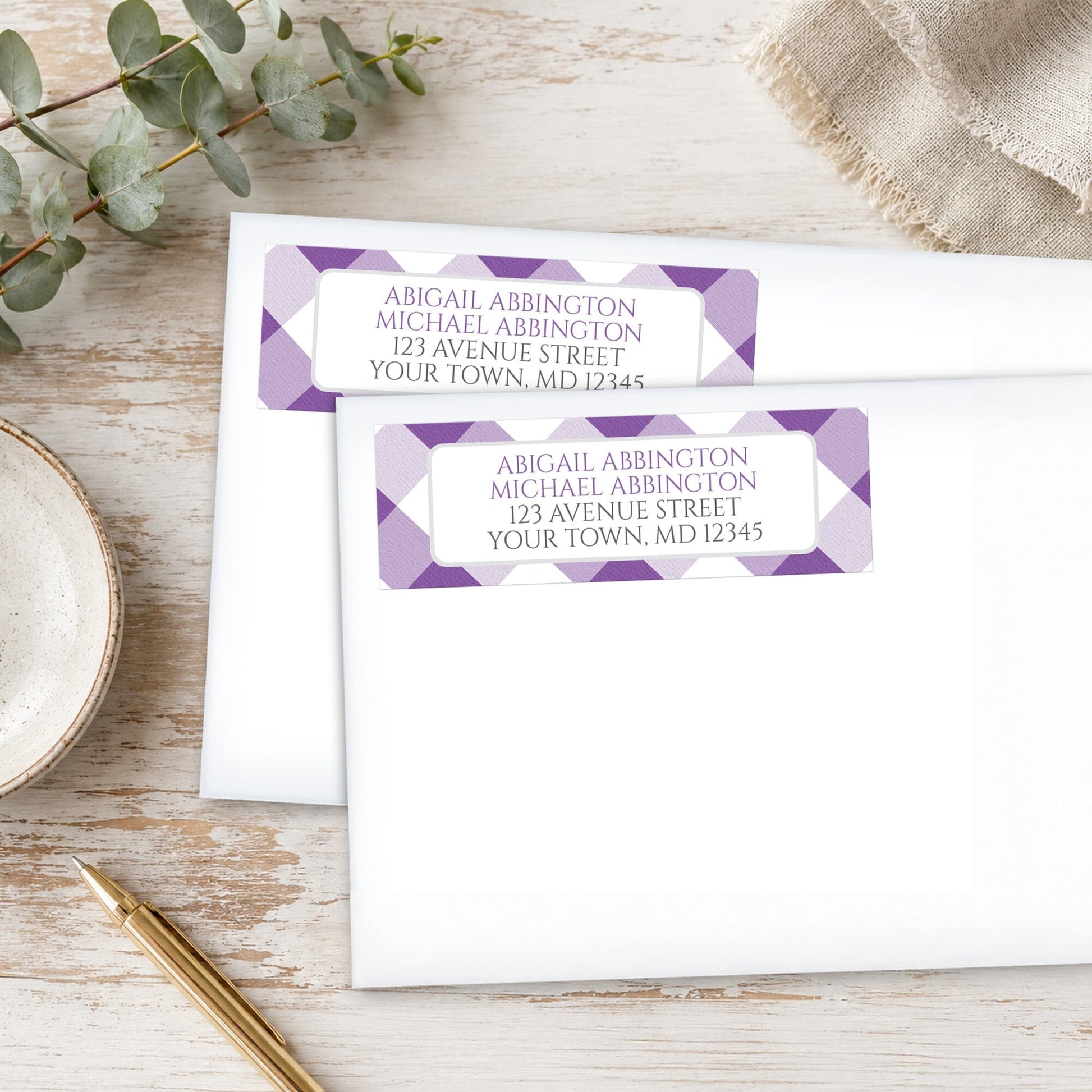 Address labels with purple gingham check pattern on white envelopes, placed on a wooden surface with eucalyptus leaves and a pen.