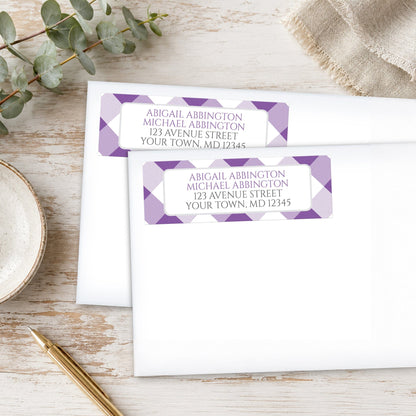 Address labels with purple gingham check pattern on white envelopes, placed on a wooden surface with eucalyptus leaves and a pen.