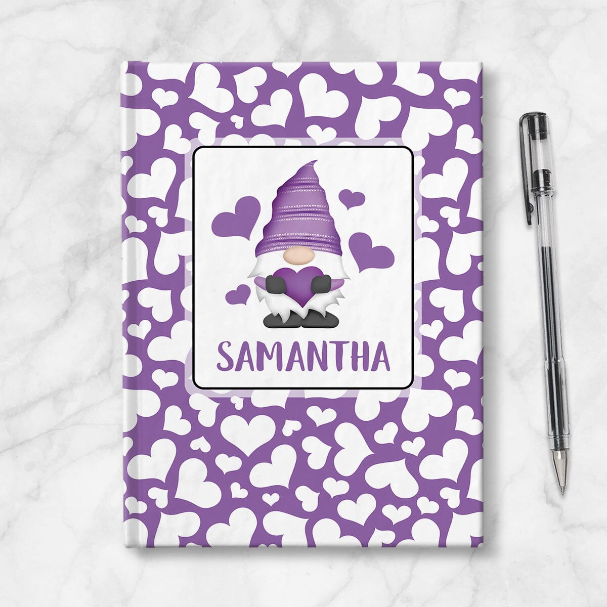Personalized Purple Hearts Gnome Journal at Artistically Invited. A custom book designed with an adorable gnome holding a purple heart with 4 hearts floating around him in a white rectangular design on the front of the book, with your name personalized in purple in a cute all capital letters font below the gnome. The background design that wraps around the cover of the book features a purple background color filled with cute multidirectional white hearts of different sizes. Image shows the book next to pen.