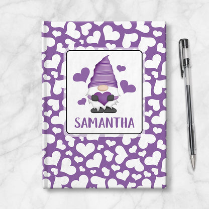 Personalized Purple Hearts Gnome Journal at Artistically Invited. A custom book designed with an adorable gnome holding a purple heart with 4 hearts floating around him in a white rectangular design on the front of the book, with your name personalized in purple in a cute all capital letters font below the gnome. The background design that wraps around the cover of the book features a purple background color filled with cute multidirectional white hearts of different sizes. Image shows the book next to pen.