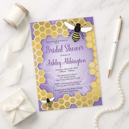 Purple Honeycomb Bee Bridal Shower Invitation on a marble surface with a pen, gift box, and pearls.