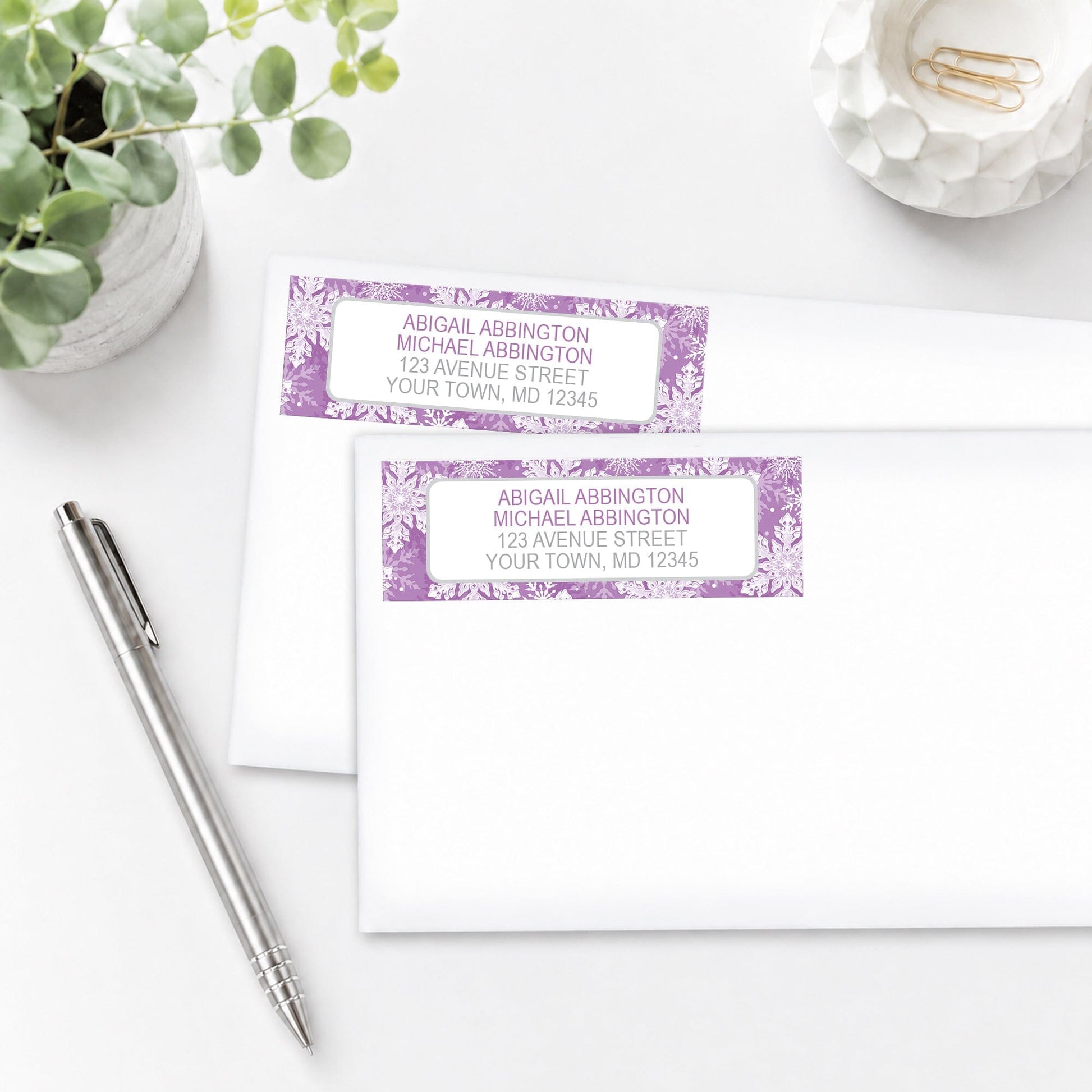 Personalized Purple Snowflake Winter Address Labels shown on white envelopes, on a white surface with a plant and a pen.