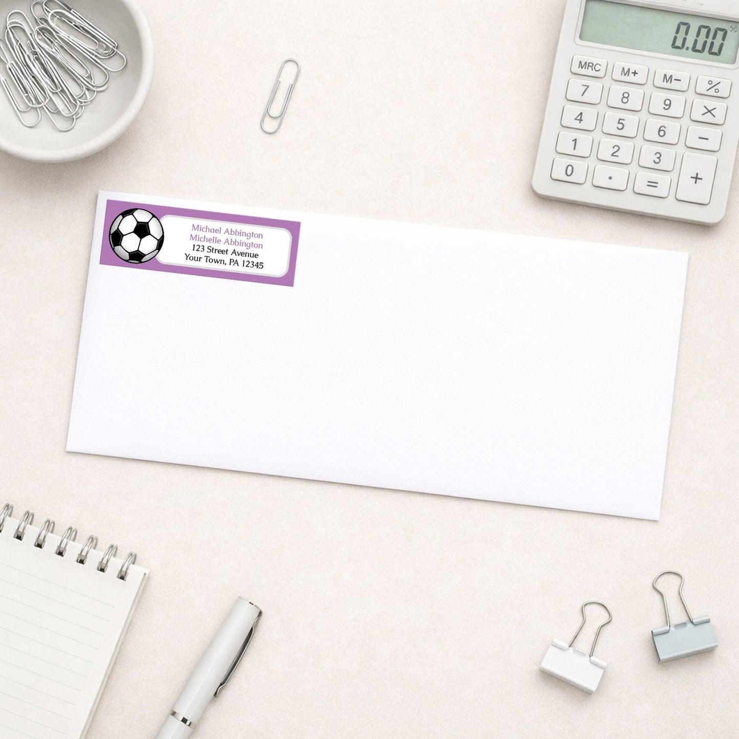 Purple Soccer Ball Return Address Label shown on a white envelope with everyday office supplies