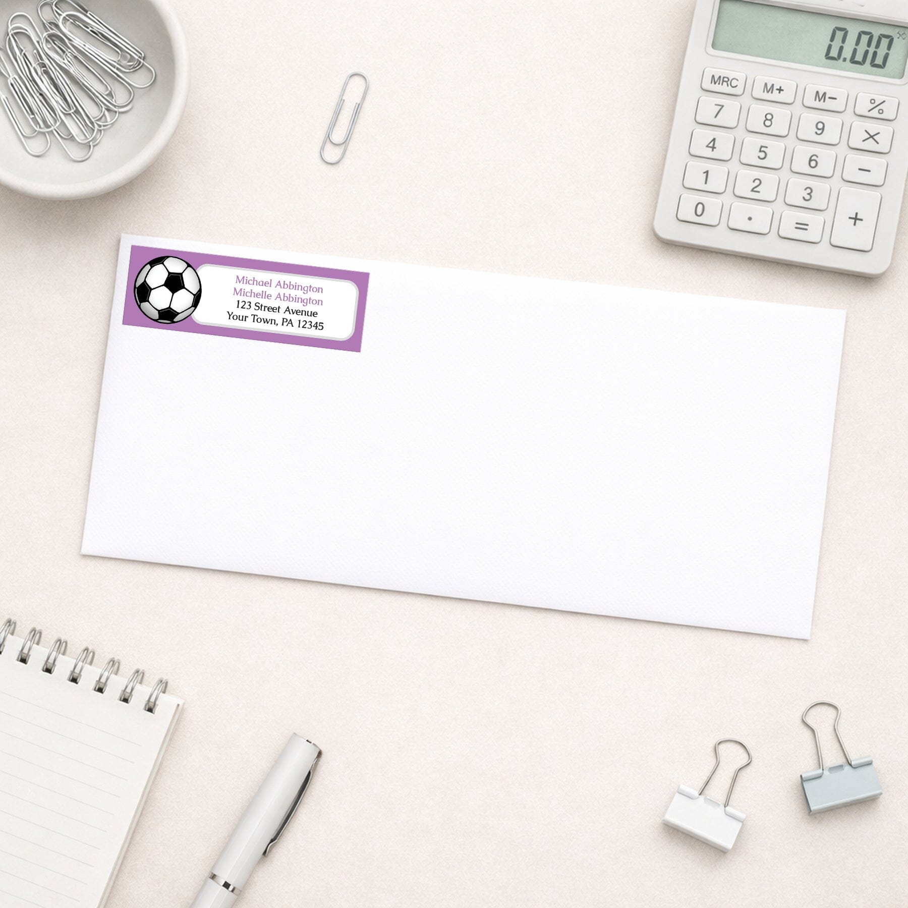 Purple Soccer Ball Return Address Label shown on a white envelope with everyday office supplies
