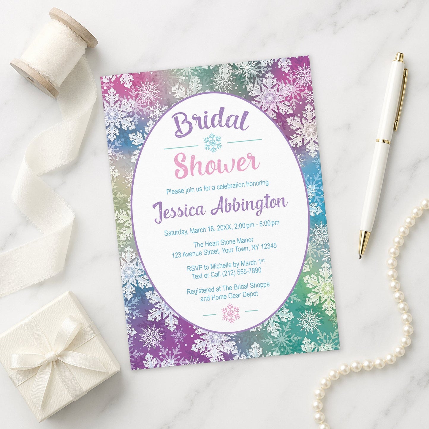 Rainbow Snowflake Bridal Shower Invitation on a marble surface with a white gift box, pen, ribbon, and pearls.