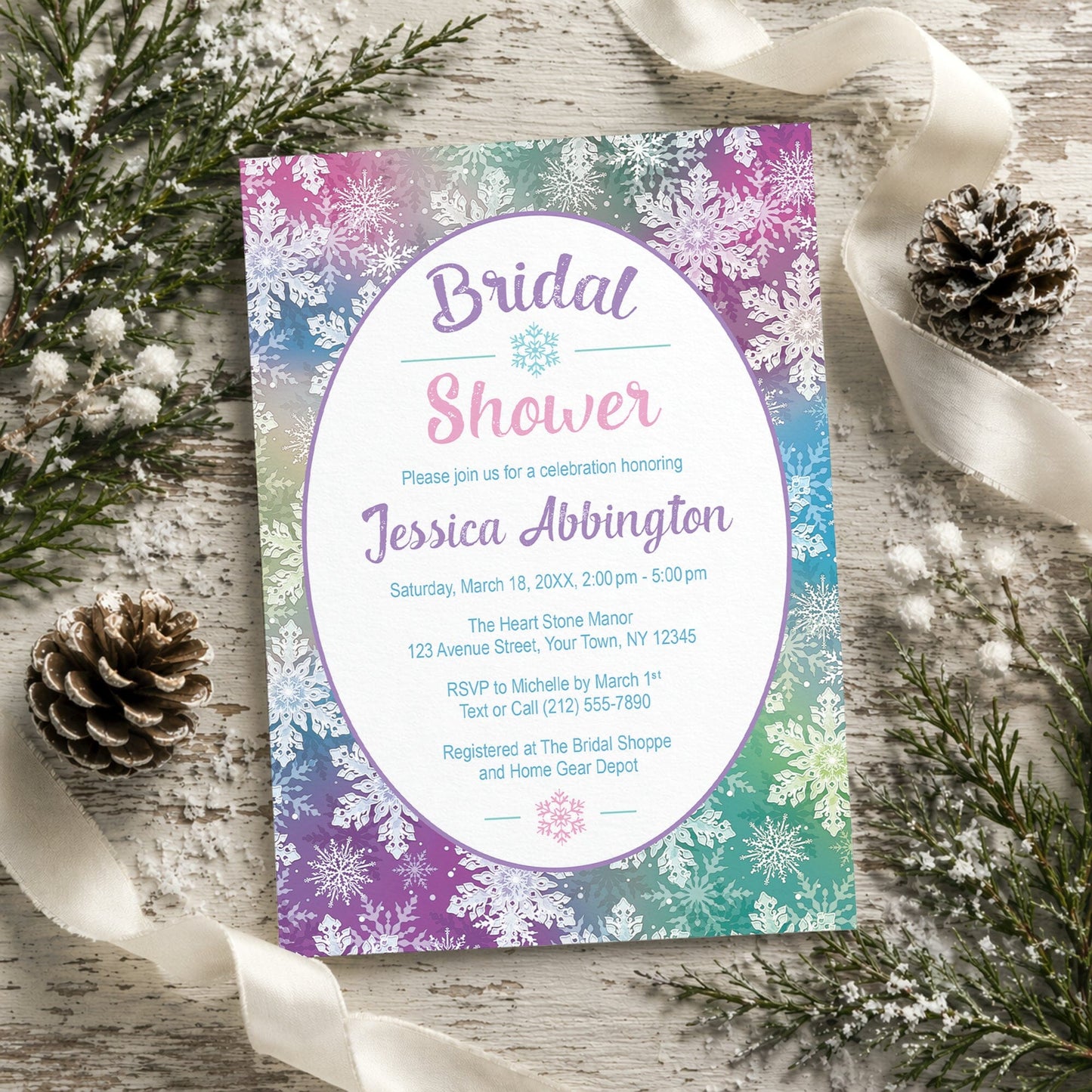 Rainbow Snowflake Bridal Shower Invitation on a rustic wooden surface with winter greenery, pine cones, and ribbon.