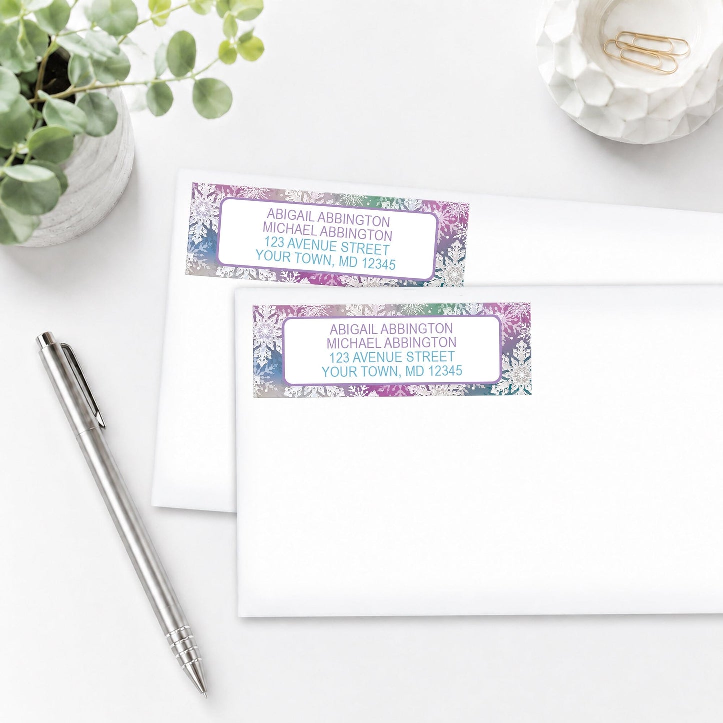 Rainbow Snowflake Winter Address Labels shown on white envelopes, on a white surface with a pen and a plant.