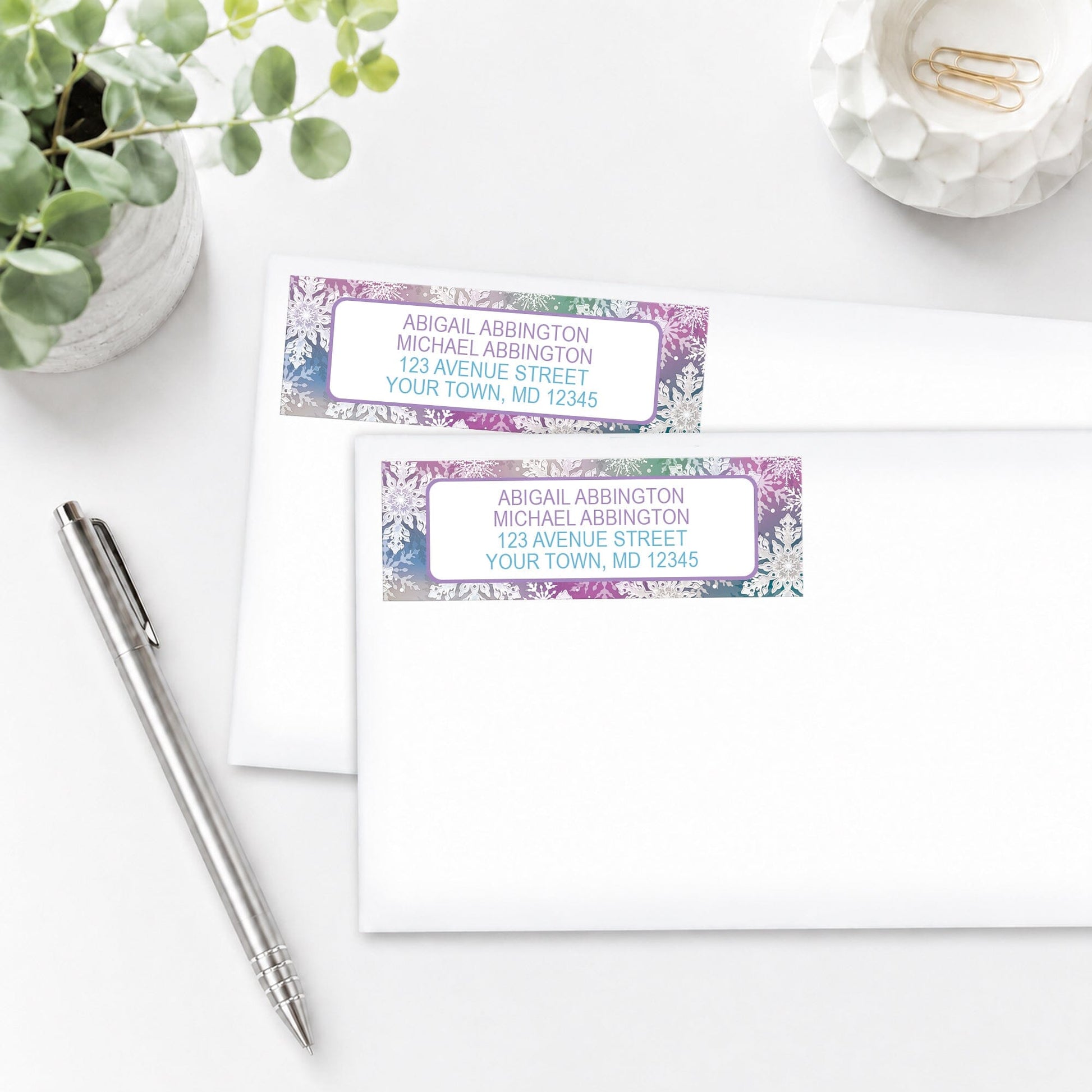 Rainbow Snowflake Winter Address Labels shown on white envelopes, on a white surface with a pen and a plant.
