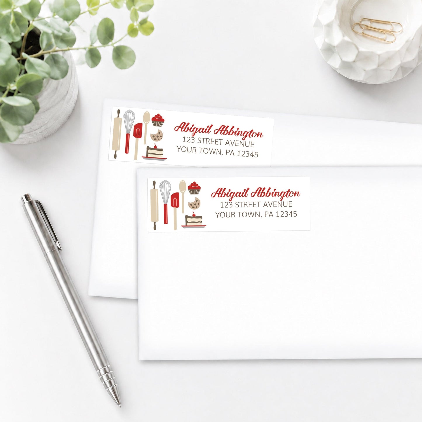 Red Baking Address Labels shown on white envelopes, on a white surface with a plant and a pen.