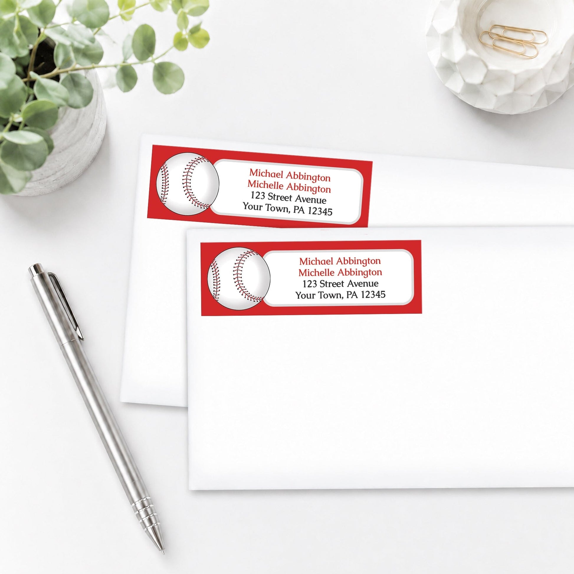 Address labels with red baseball design on envelopes with a pen and plant in the background.