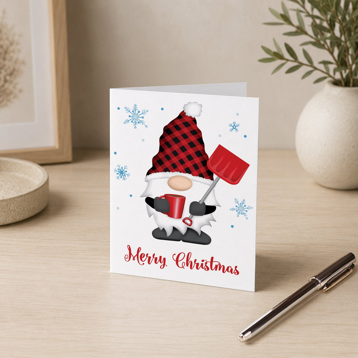 Folded Christmas card with white-bearded gnome wearing a red and black buffalo plaid hat, holding a red snow shovel and mug, blue snowflakes, and Merry Christmas lettering, displayed on a wooden desk with a pen and home decor in the background.