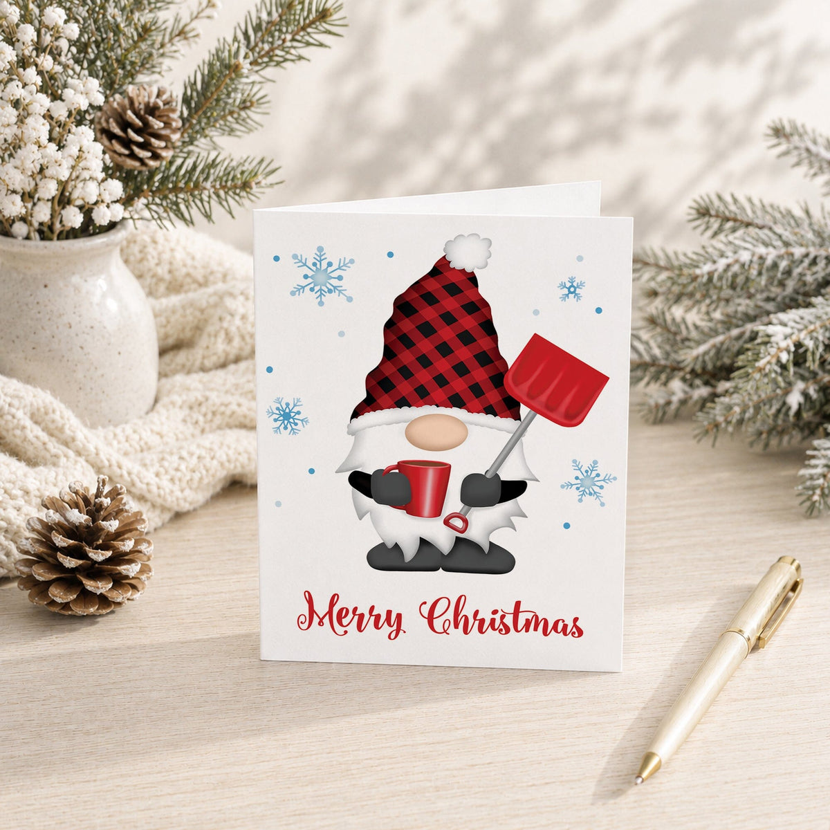 Folded Christmas card with white-bearded gnome wearing a red and black buffalo plaid hat, holding a red snow shovel and mug, blue snowflakes, and Merry Christmas lettering, shown on a light desk with winter greenery, a vase, a pine cone, and a pen.