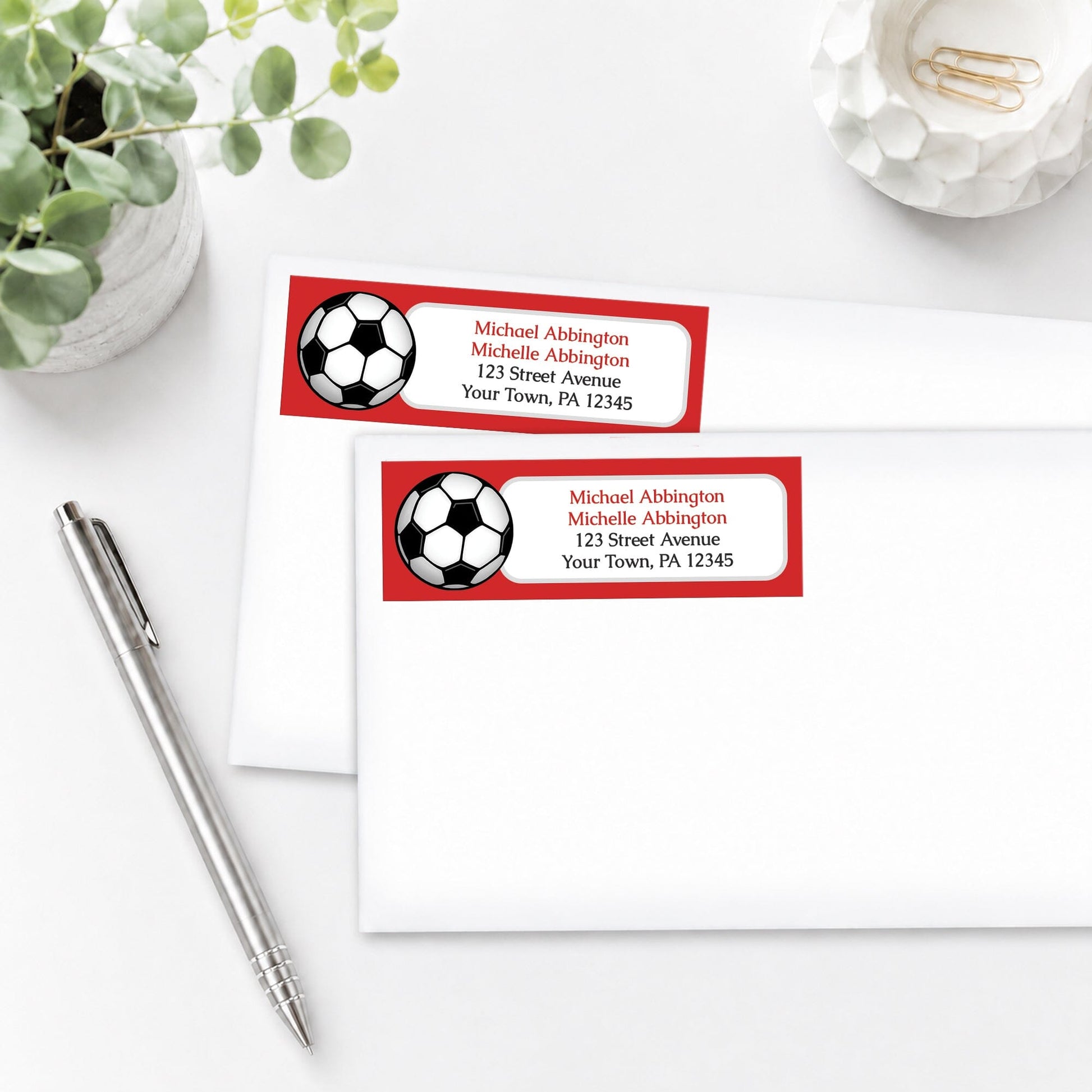 Red Soccer Ball Address Labels on white envelopes, shown on a white surface with a plant and a pen.