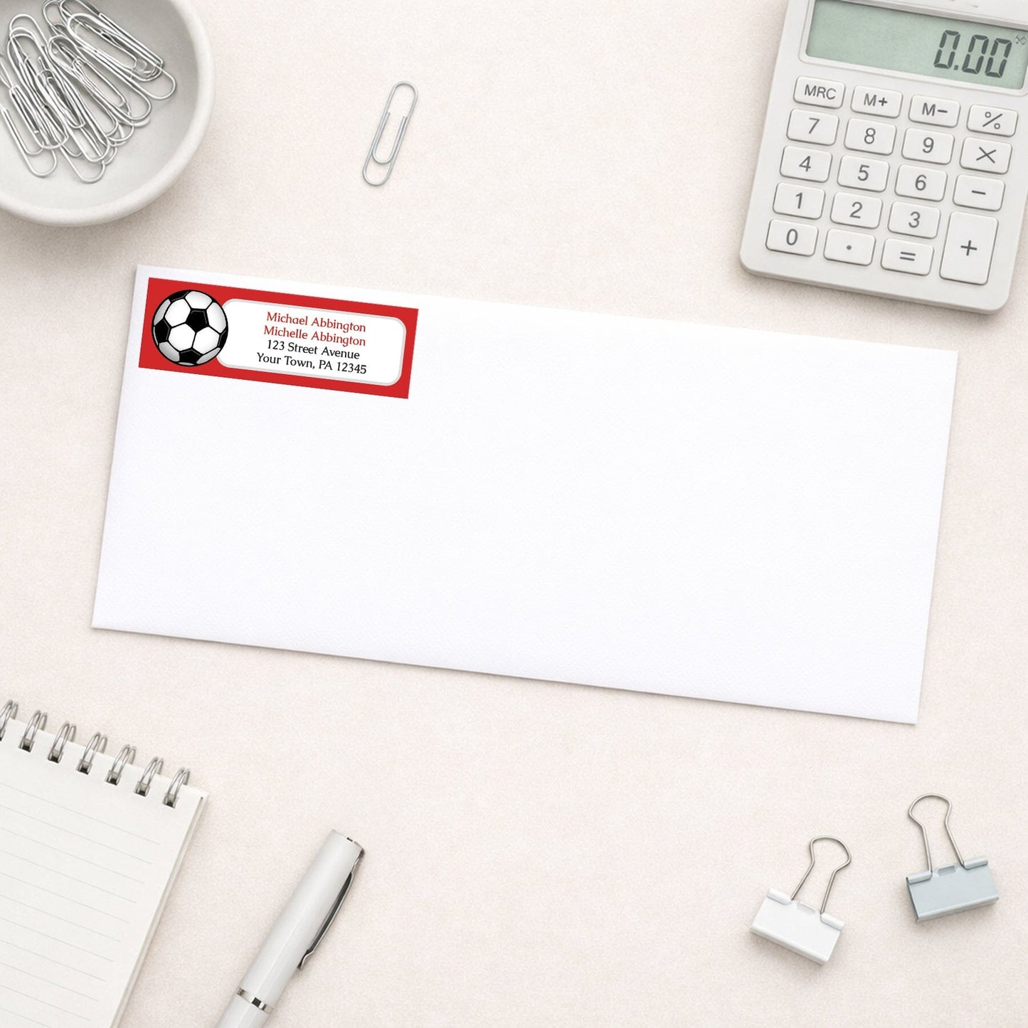 Red Soccer Ball Return Address Label shown on a white envelope with everyday office supplies