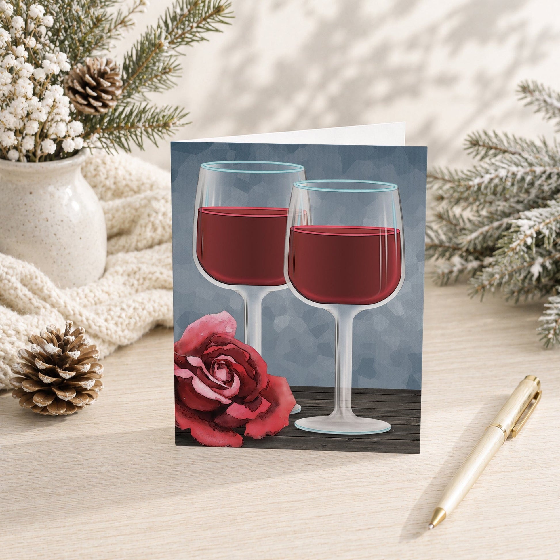 Red Wine Glasses Floral Rose Note Card shown on a tabletop with a vase, winter greenery, pine cone, and a pen.