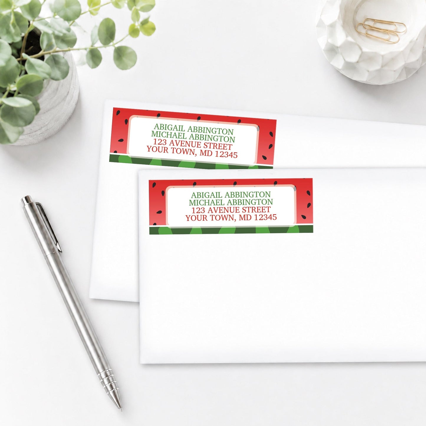 Red and Green Watermelon Address Labels, shown on white envelopes, on a white surface with a plant and a pen.