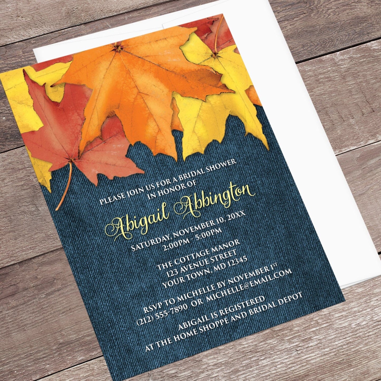 Rustic Autumn Leaves Denim Bridal Shower Invitation shown at an angle on a wooden surface with a white envelope.