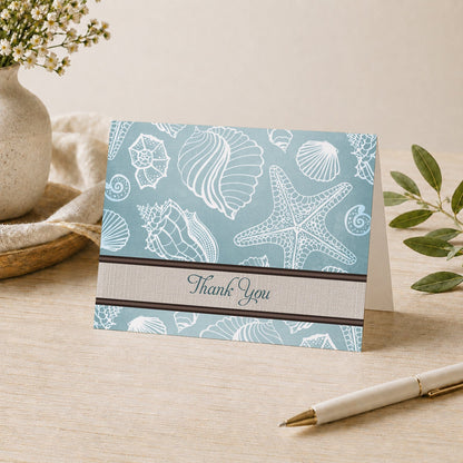 Rustic Beach Linen Seashell Thank You Card shown on a desk with a vase, plants, and a pen.