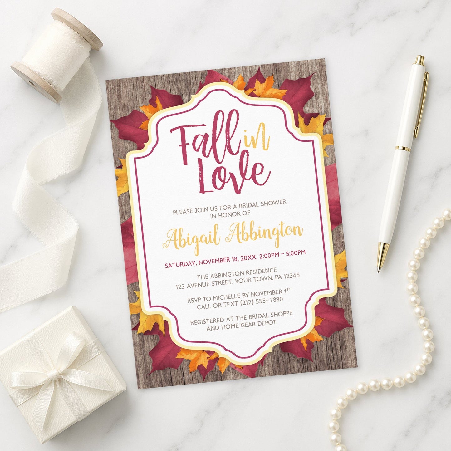 Rustic Burgundy Gold Bridal Shower Invitation with 'Fall in Love' theme on a marble surface with decorative items.