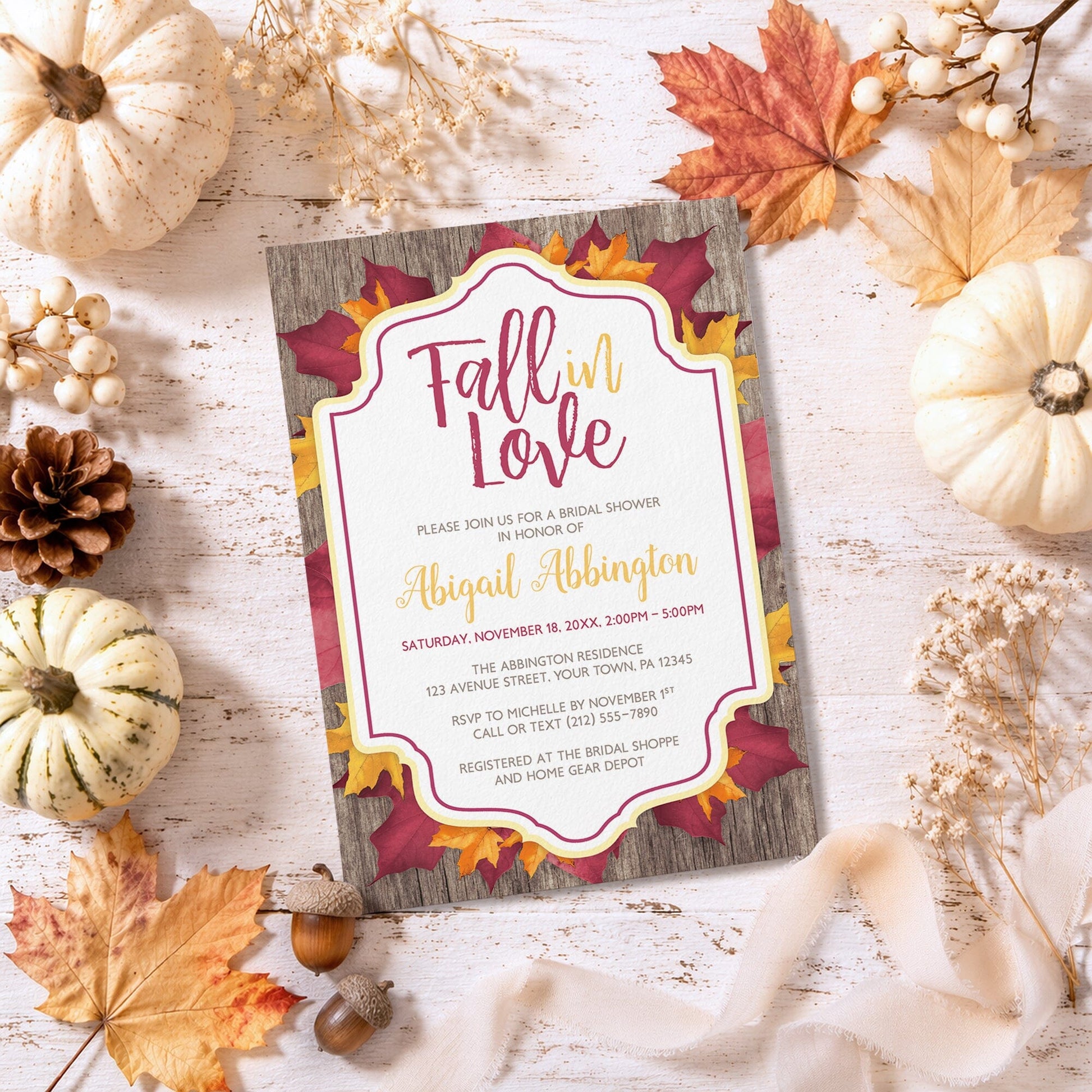 Rustic Burgundy Gold Bridal Shower Invitation with 'Fall in Love' theme surrounded by autumn pumpkins, leaves, and pinecones on a wooden surface.