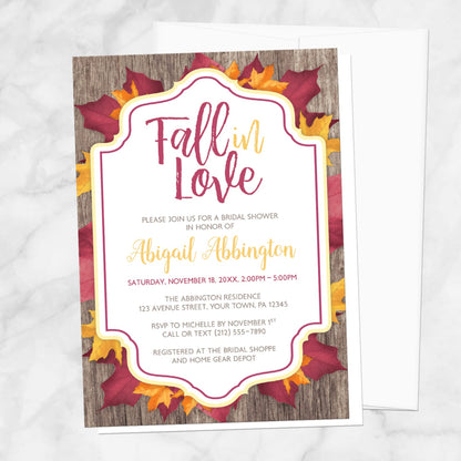 Fall bridal shower invitation with burgundy and gold-colored leaves and rustic wood with a Fall in Love title, displayed with a white envelope on a marble surface.
