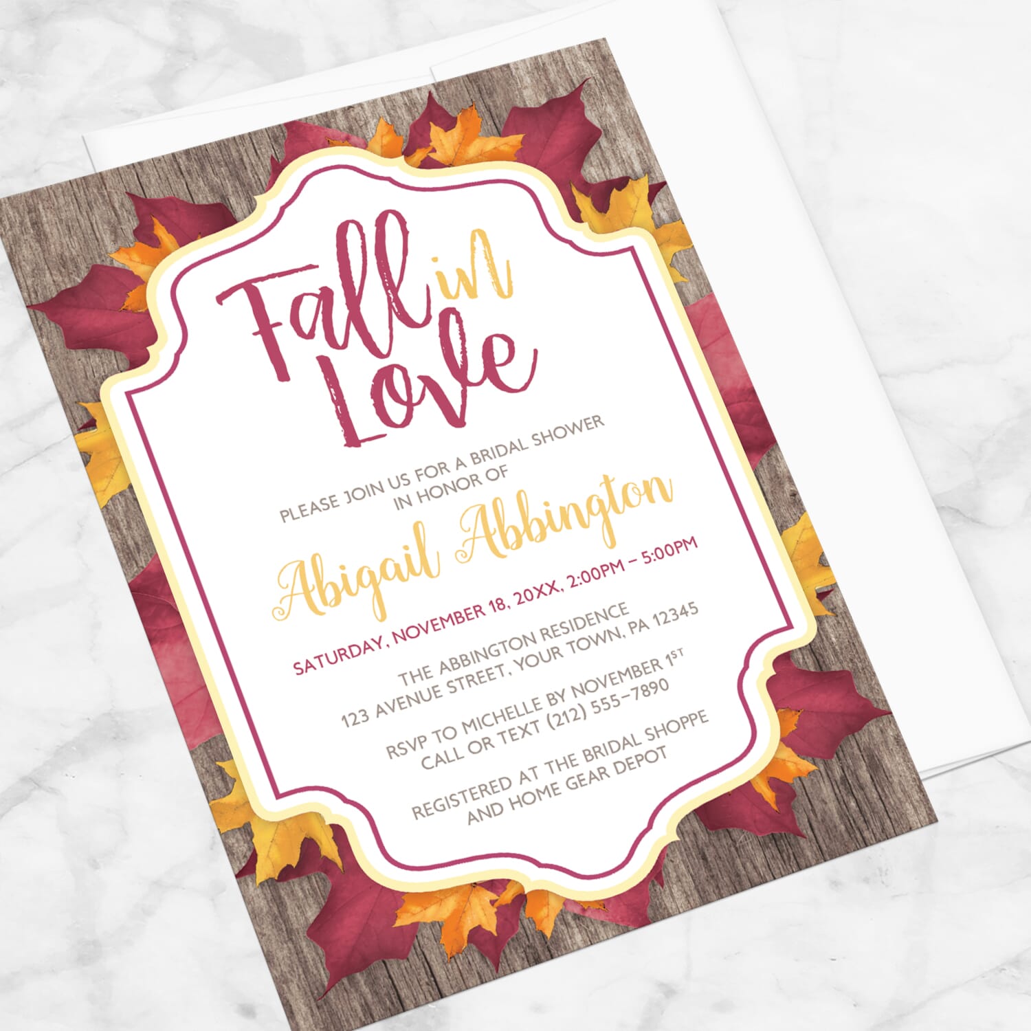 Fall bridal shower invitation with burgundy and gold-colored leaves and rustic wood with a Fall in Love title, displayed at an angle with a white envelope on a marble surface.