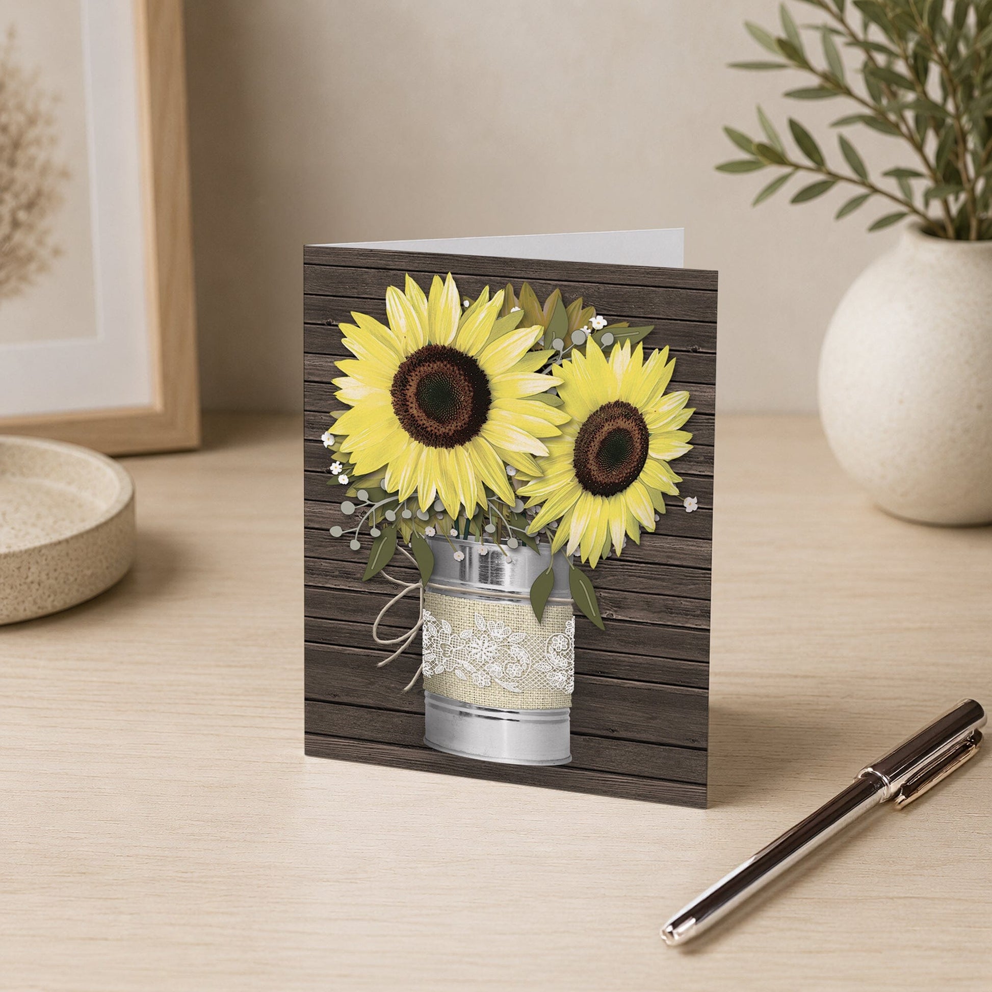Rustic Burlap and Lace Tin Can Sunflower Note Card shown on a desk with a plant and a pen.