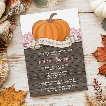 A fall-themed baby shower invitation featuring a large orange pumpkin illustration, pink lace, an "a little Pumpkin is on her way" banner, and rustic wood-textured background, styled on a whitewashed wooden surface with autumn leaves, a knitted blanket, and small pumpkins.