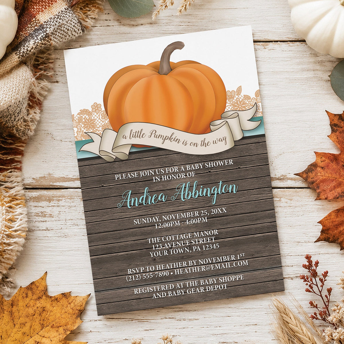 Rustic pumpkin baby shower invitation with orange pumpkin, teal script name, white uppercase details, illustrated ribbon, and wood plank background, on a wooden tabletop with autumn leaves, fall pumpkins, and a cozy blanket beside it.