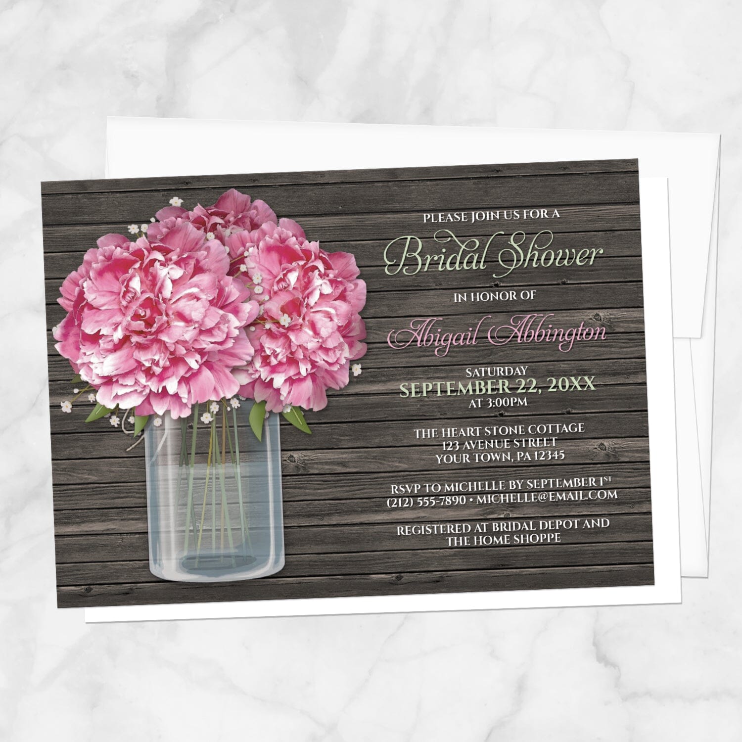 Bridal shower invitations with pink peony flowers in a glass mason jar over a dark wood background, displayed with a white envelope on a marble surface