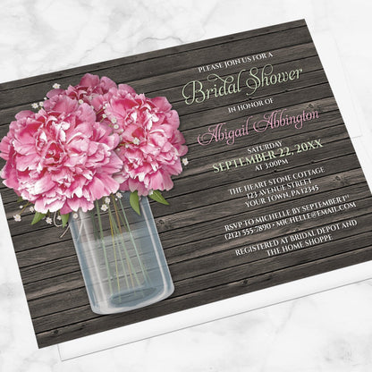 Bridal shower invitations with pink peony flowers in a glass mason jar over a dark wood background, displayed at an angle with a white envelope on a marble surface